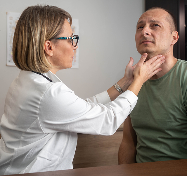 Female doctor with short blonde hair wearing a white coat and checking for thyroid nodules on male patient's neck. Male patient is wearing a green short sleeved t-shirt. doctor checking thyroid nodules