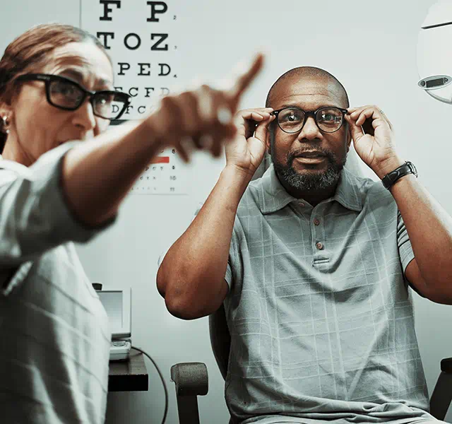 Older man getting an eye exam. He is wearing a gray polo T-shirt and holding glasses up to his eyes while a doctor points at something for him to see. dummy mobile image