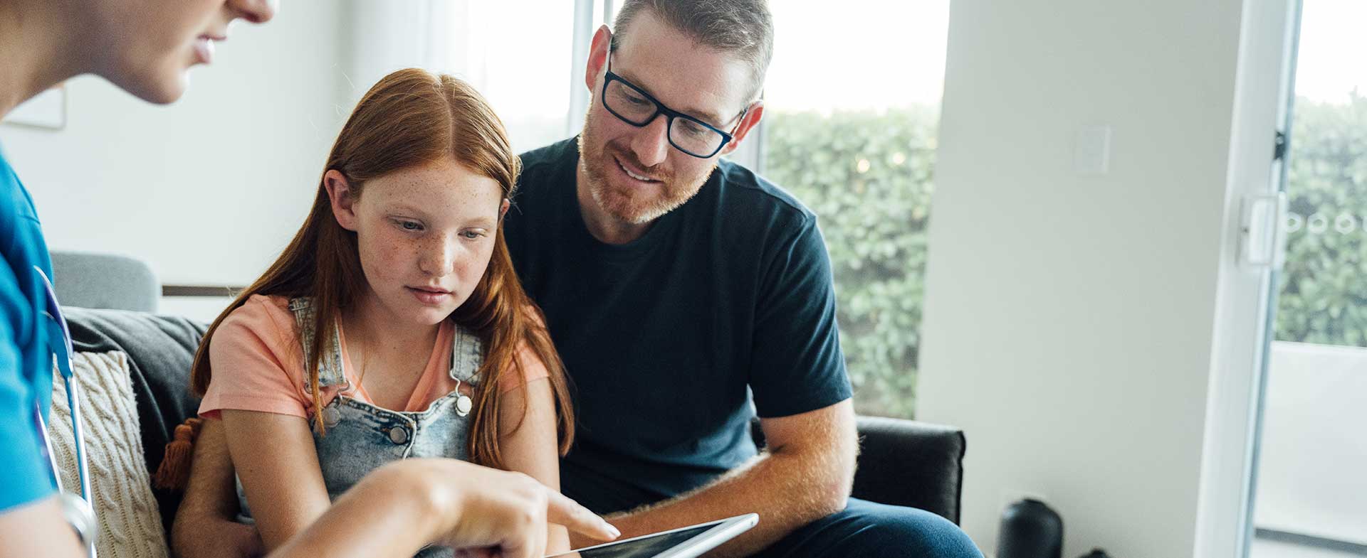 A father with facial hair and glasses sits on a couch with his pre-teen redhead daughter. They are sitting with a nurse in a blue top who is reviewing information with them on a tablet. autism healthcare transition