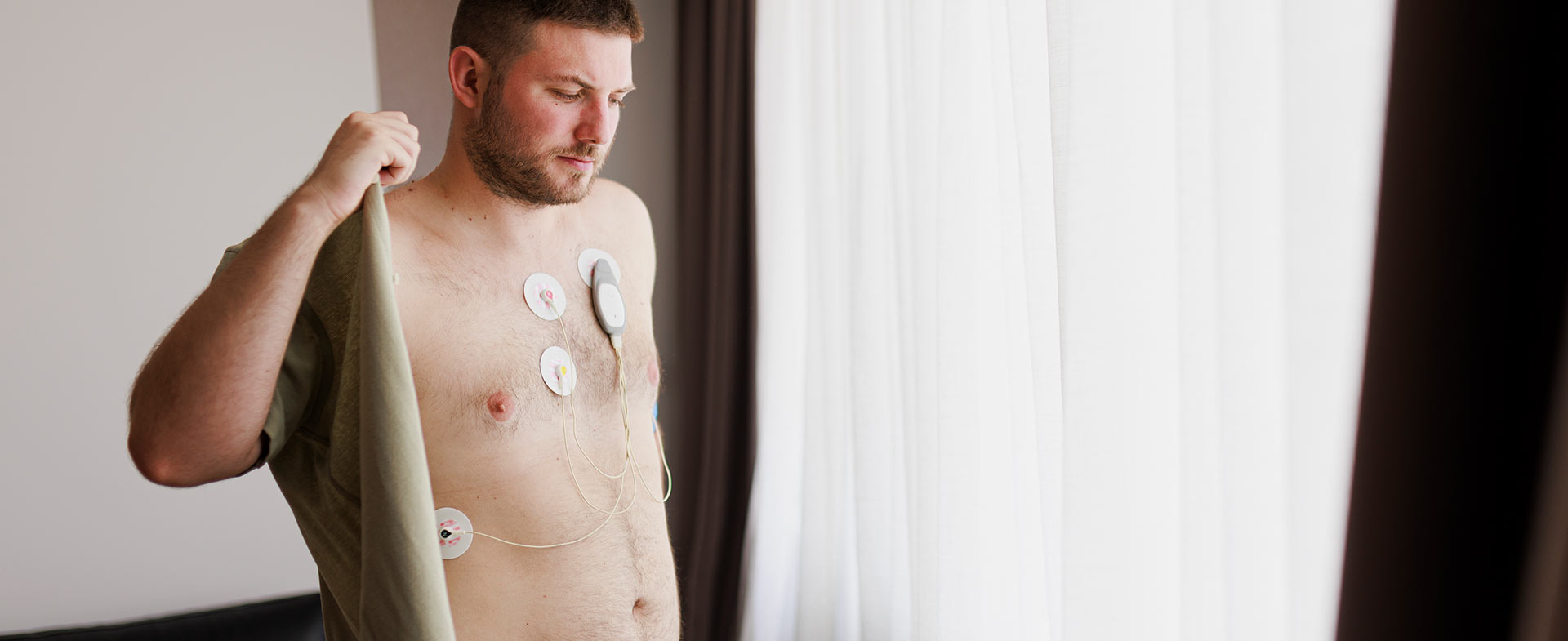 A man with medical electrodes attached to his chest standing near a window in a softly lit room while putting on a button down shirt. The background includes a section of a sofa with cushions. heart monitors