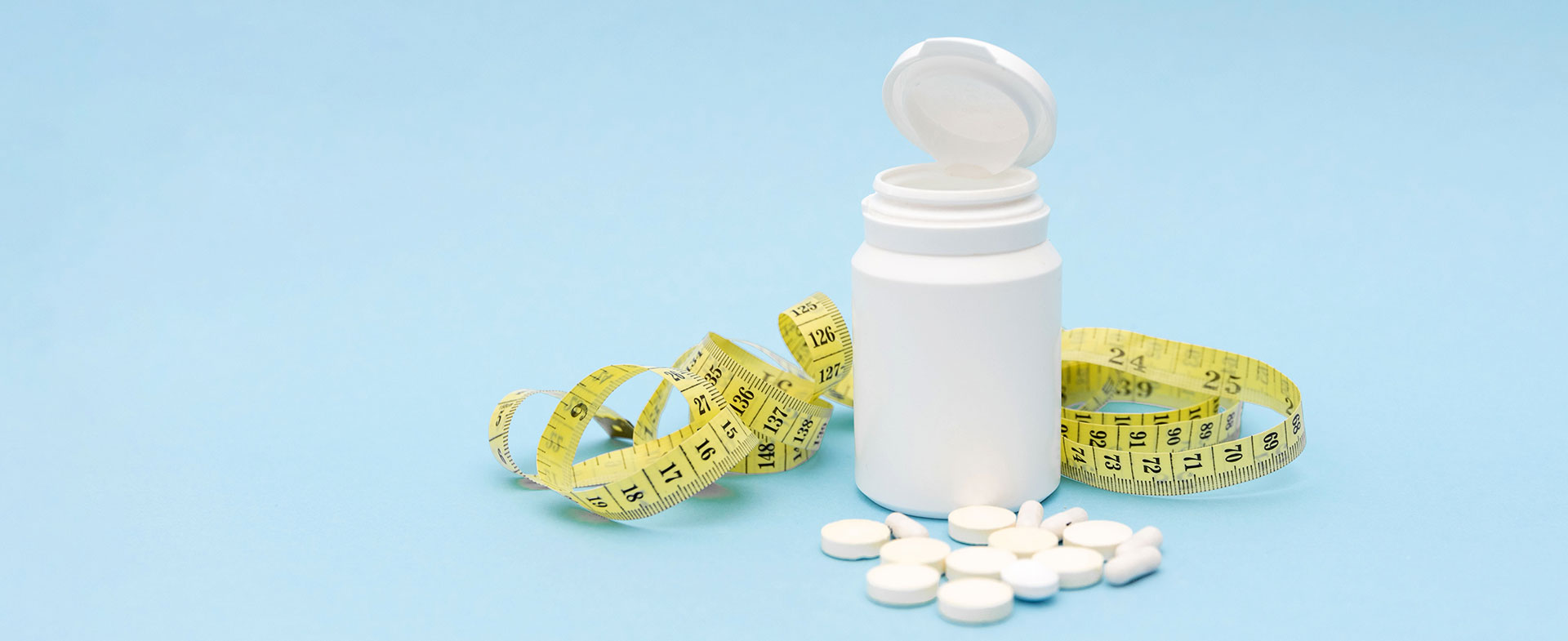 A white bottle with an open lid is on a light blue background, surrounded by a yellow measuring tape and several white pills scattered around the bottle. next generation weight loss meds
