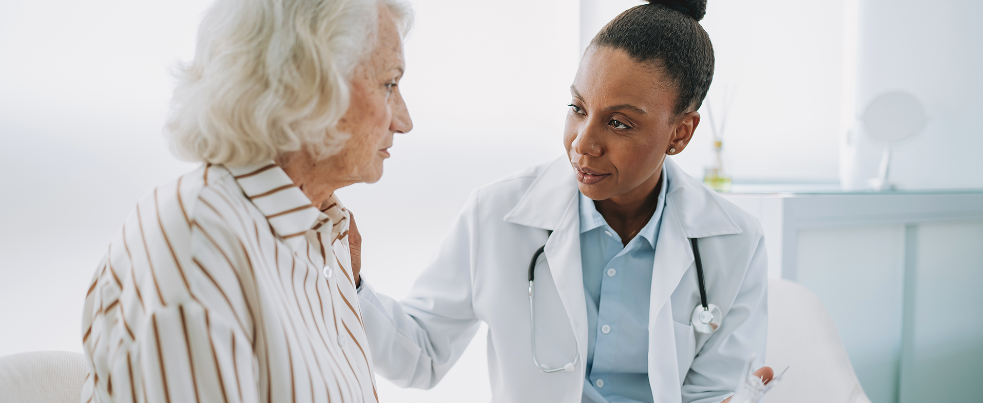 Older woman with white hair and white button-down shirt with vertical brown stripes talking to female doctor wearing a white coat, blue button-down shirt and hair in a bun. doctor talking to patient