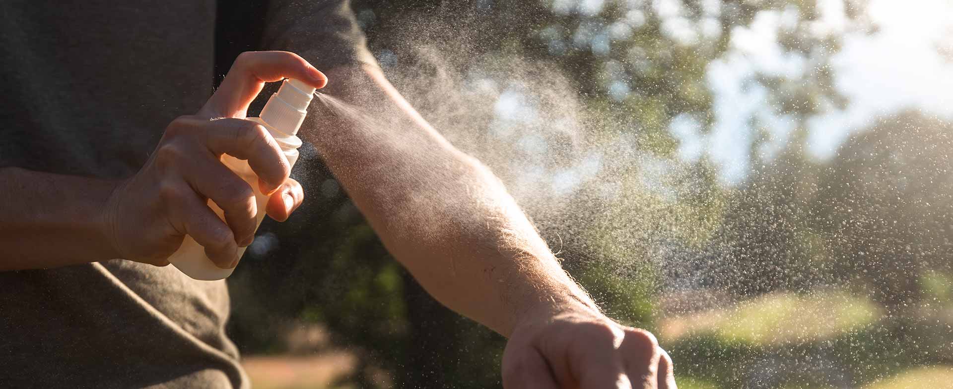 A person outdoors sprays insect repellent onto their arm, with sunlight filtering through trees in the background. repelling mosquitoes