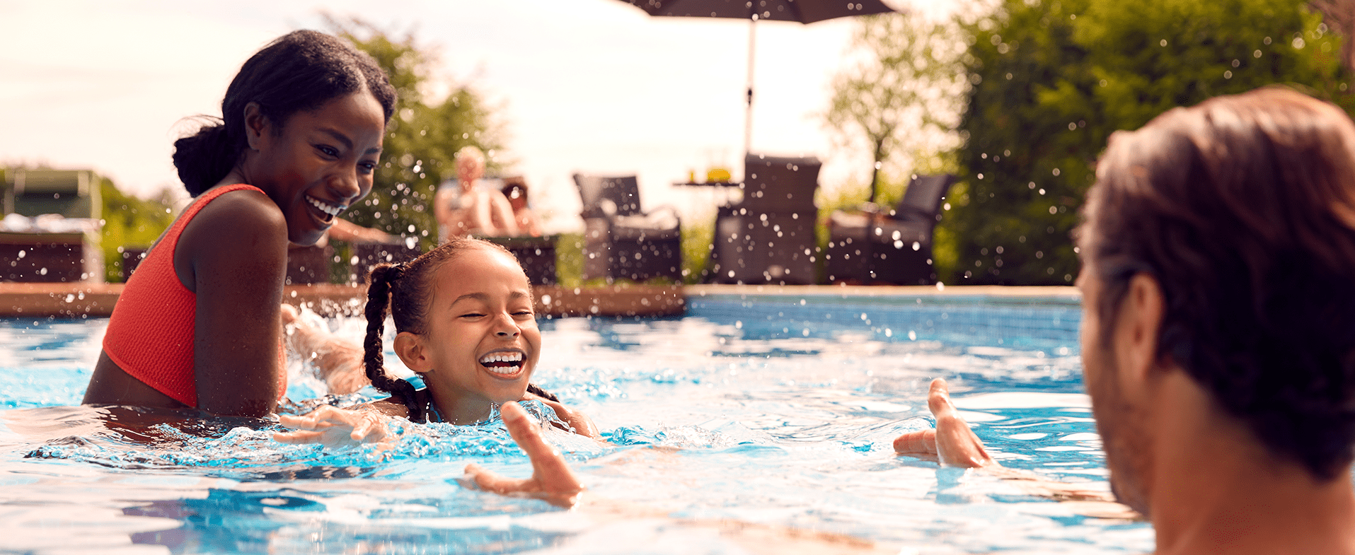 A family of three swimming outside in a pool. The mom is wearing a neon pink two-piece bathing suit and smiling, the daughter has braids in her hair and is smiling and the dad has his arms opened out to his smiling child. dummy desktop Image