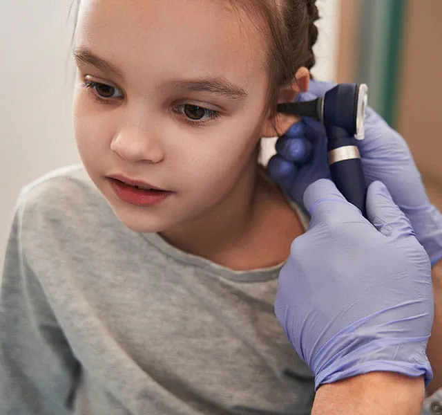 A person wearing blue gloves examines a young girl's ear with an otoscope in a medical setting. kid ear infections