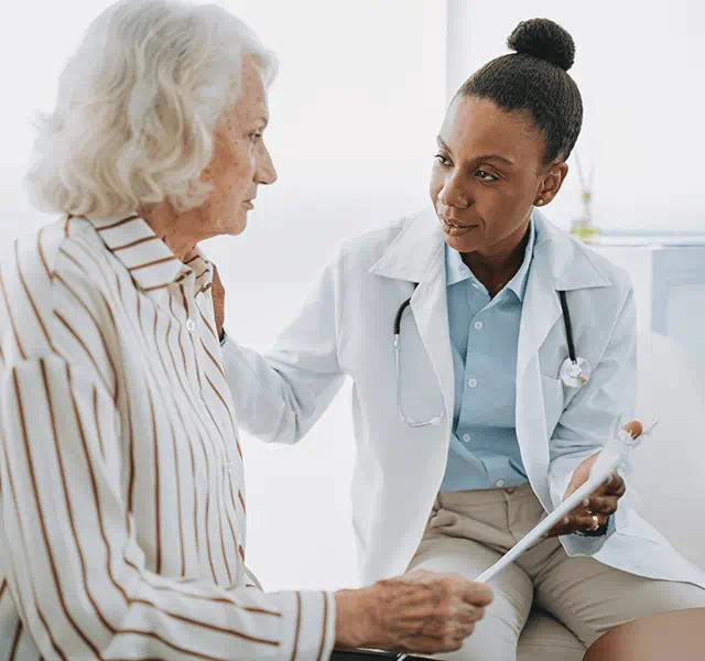 Older woman with white hair and white button-down shirt with vertical brown stripes talking to female doctor wearing a white coat, blue button-down shirt and hair in a bun. doctor talking to patient