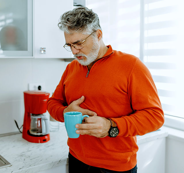 A man with a gray beard wearing an orange sweater stands in a kitchen holding a blue mug. He appears to be experiencing discomfort, with one hand on their chest. A red coffee maker and various kitchen items are on the countertop nearby. gerd