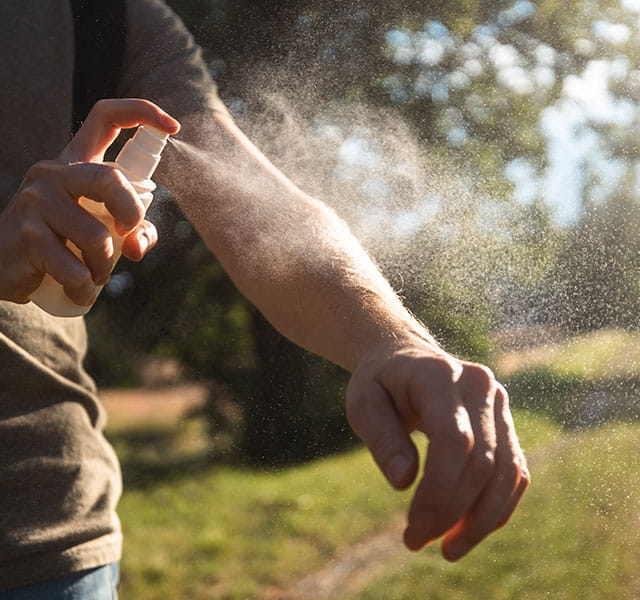 A person outdoors sprays insect repellent onto their arm, with sunlight filtering through trees in the background. repelling mosquitoes