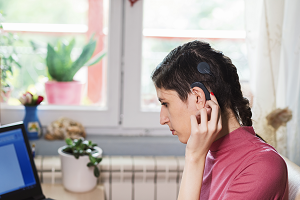 A woman adjusts her cochlear implant while sitting in front of a computer screen.